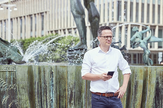 Per Stigenberg 3 står framför en fontän och håller i en mobiltelefon.