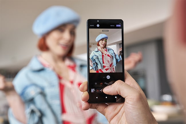 Närbild på person som fotograferar kvinna i basker, mobilskärm i förgrunden och kvinnan i blurrad bakgrund.